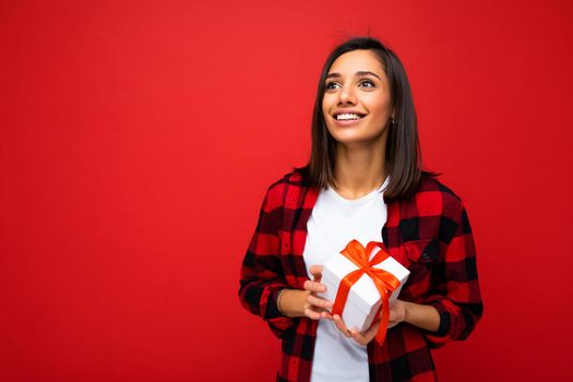 Shot of attractive positive smiling young brunette woman isolated over colourful background wall wearing everyday trendy outfit holding gift box and looking up to the side. Copy space