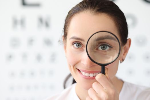 Woman optometrist holding magnifying glass in eyes against background of vision examination table. Correction of myopia concept