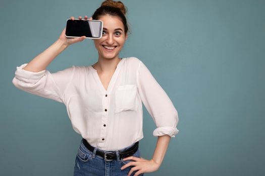 Photo of beautiful smiling young woman good looking wearing casual stylish outfit standing isolated on background with copy space holding smartphone showing phone in hand with empty screen display for mockup looking at camera.