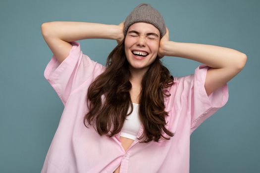 Photo of beautiful emotional young brunette woman standing isolated over blue background wall wearing pink shirt and gray hat and having fun.