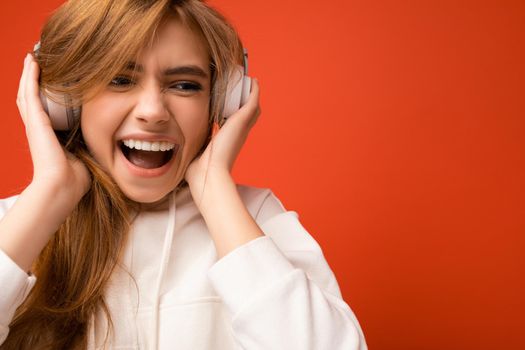 Portrait photo shot of attractive emotional positive young blonde woman wearing white hoodie isolated over colourful background wearing white wireless bluetooth headsets listening to music and having fun.