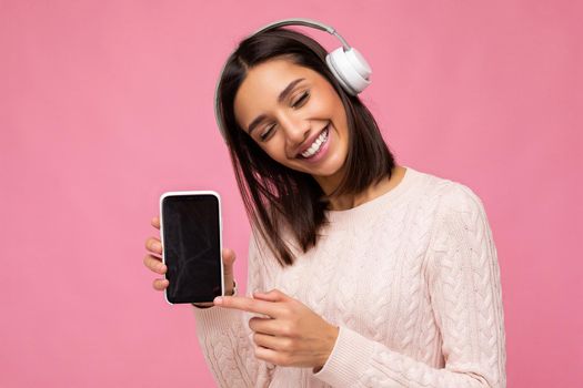 Beautiful cute young brunette woman wearing pink sweater isolated over pink background wall wearing white bluetooth wireless headphones and listening to good music and showing mobile phone with empty display for mockup having fun.