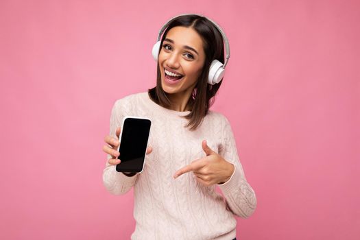 Attractive positive smiling young woman wearing stylish casual outfit isolated on colourful background wall holding and showing mobile phone with empty screen for cutout wearing white bluetooth headphones and having fun looking at camera and pointing finger at gadjet display.