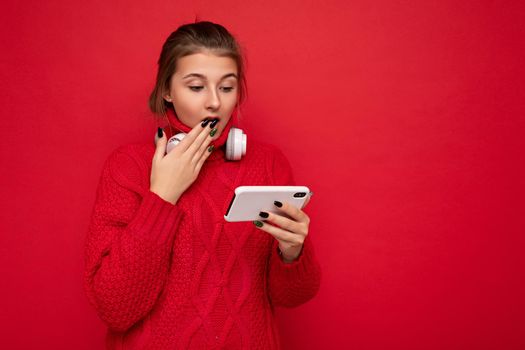 Attractive shocked young brunet woman wearing red sweater isolated over red background wall holding and using mobile phone surfing on the internet and wearing bluetooth headphones looking at smartphone. empty space