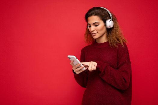 Shot of beautiful young brunette curly woman wearing dark red sweater isolated over red background wall wearing white wireless headphones and listening to music and using mobile phone surfing on the internet looking at smartphone display. copy space