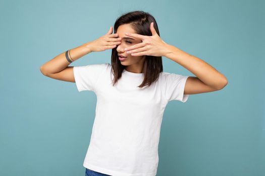 Portrait of young beautiful brunette woman with sincere emotions wearing casual white t-shirt for mockup isolated on blue background with empty space and covering eyes with hands.