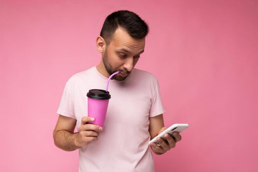 Handsome happy concentrated young brunette unshaven man with beard wearing white t-shirt isolated over pink backgroung wall using mobile phone holding paper coffee cup for mock up and drinking looking at gadjet display. copy space