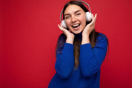 Attractive positive young brunette woman wearing blue sweater isolated over red background wall wearing white bluetooth earphones listening to cool music and having fun looking at camera.