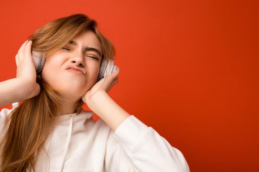 Portrait photo shot of attractive emotional positive young blonde woman wearing white hoodie isolated over colourful background wearing white wireless bluetooth headsets listening to music and enjoying having fun.