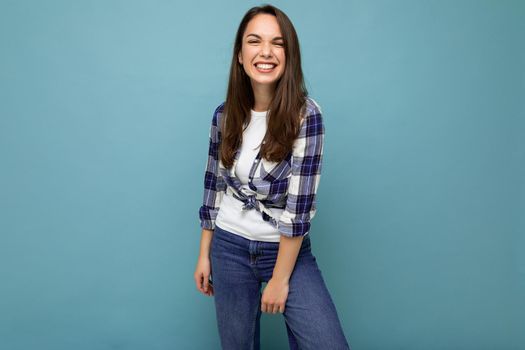 Photo portrait of young beautiful smiling hipster brunette woman in trendy blue and white shirt and jeans clothes. Sexy carefree female person posing isolated near blue wall with empty space in studio. Positive model with natural makeup.