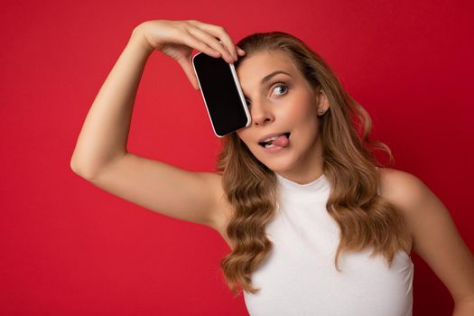 Happy sexy attractive young blonde female person wearing white t-shirt isolated on red background with copy space holding smartphone showing phone in hand with empty screen for cutout looking to the side and showing tongue.
