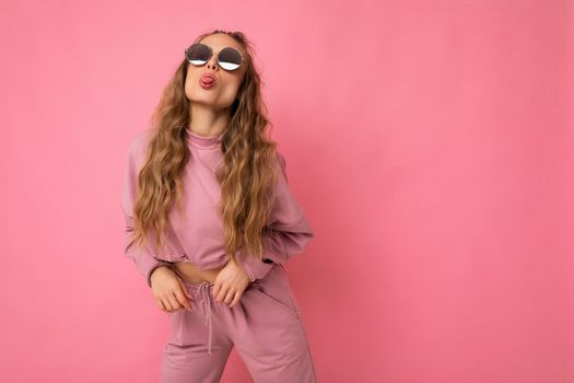 Shot of fascinating positive happy young dark blonde curly woman isolated over pink background wall wearing casual pink sport clothes and stylish sunglasses and having fun. empty space
