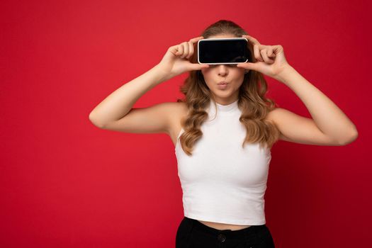 funny young blonde woman wearing white t-shirt isolated on red background with copy space holding smartphone showing phone in hand with empty screen for cutout.