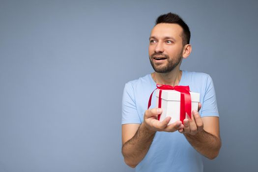 Shot of handsome joyful brunette unshaven young man isolated over blue background wall wearing blue t-shirt holding white gift box with red ribbon and looking to the side. Copy space