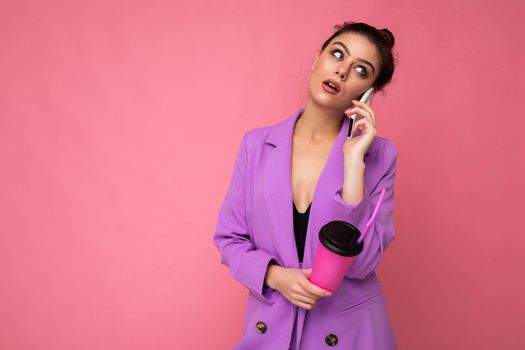 Portrait of pretty brunette woman wear trendy purple jacket, talking on smart phone, holding takeaway coffee in paper cup, isolated over pink background blank space for advertising. Empty space