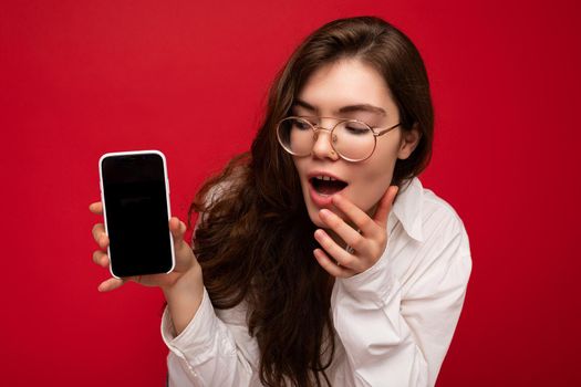 photo of sexy amazed beautiful young brunette woman wearing white shirt and optical glasses isolated over red background holding in hand and showing mobile phone with empty screen for cutout looking at gadjet screen.