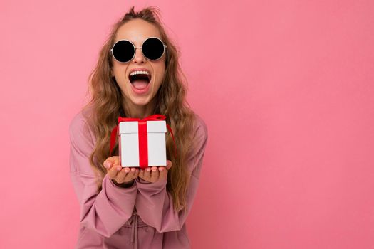 Photo shot of pretty positive surprised young blond woman isolated over colourful background wall wearing trendy outfit look holding gift box and looking at camera. Copy space, mockup