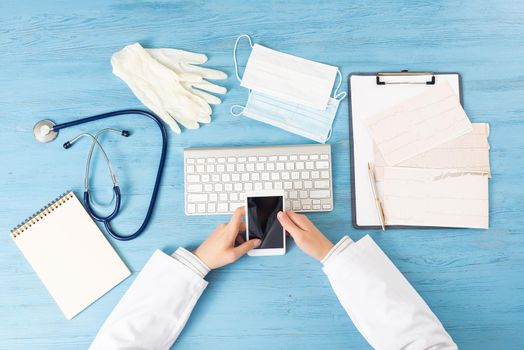 Top view of doctor hand holding smartphone. Cardiac examination in hospital. Therapist sitting at blue wooden desk with stethoscope, medical gloves and mask. Examination and consultation in clinic