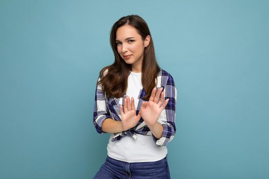 Young serious dissatisfied beautiful brunette woman with sincere emotions wearing trendy check shirt standing isolated on blue background with empty space and showing stop gesture saying no. Negative concept. I don't want it.