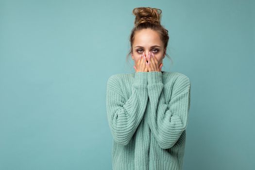 Portrait of young shocked nice winsome blond woman with sincere emotions wearing trendy blue sweater isolated over blue background with free space and covering mouth.