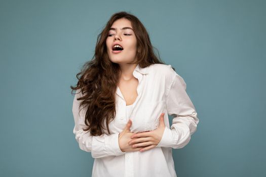 Portrait of young emotional shocked beautiful wave-haired brunette woman with sincere emotions wearing casual white shirt isolated on blue background with empty space and touches chest with hands.