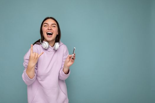 Photo of beautiful positive smiling young brunet woman wearing purple hoodie isolated on blue background holding and using smartphone wearing white wireless earphones having fun and showing rock and roll gesture.