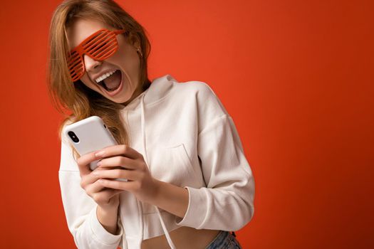 Closeup photo of shocked emotional young blonde woman wearing stylish white hoodie and funny colorful glasses standing isolated over colourful background holding and using mobile phone communicating online on the internet looking at gadjet and shouting.