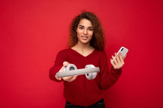 Charming funny young brunette curly woman wearing dark red sweater isolated on red backdrop holding and suggesting white wireless headphones and mobile phone looking at camera.