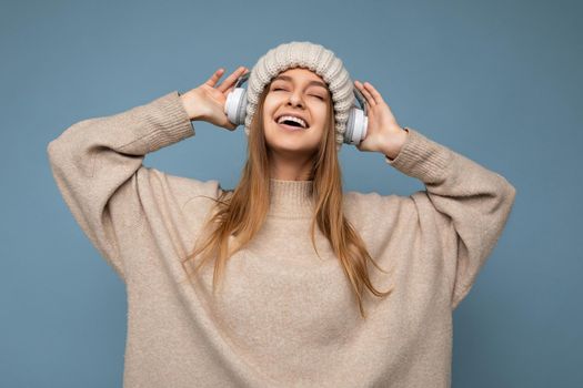 Photo of beautiful happy smiling young blonde woman wearing beige winter sweater and hat isolated over blue background wearing white wireless bluetooth headphones listening to cool music enjoying with close eyes.