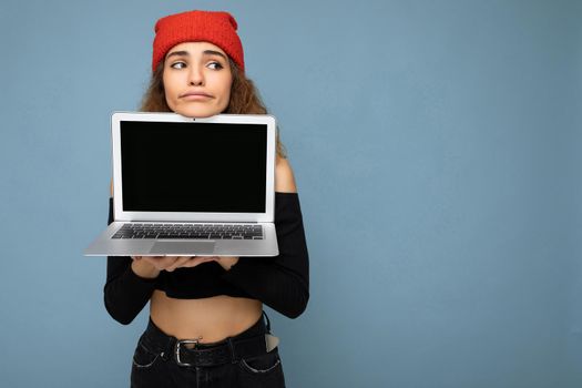 Beautiful funny amusing young brunet curly girl wearing black crop top and red and orange do-rag isolated over light blue wall background holding computer laptop with empty screen monitor looking to the side and having doubts. Mock up, copy space, empty space