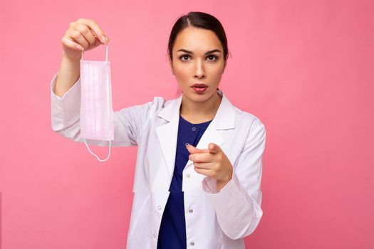 Caucasian woman showing protection face mask isolated on pink background and pointing finger at camera. Coronavirus or Covid-19 Concept.