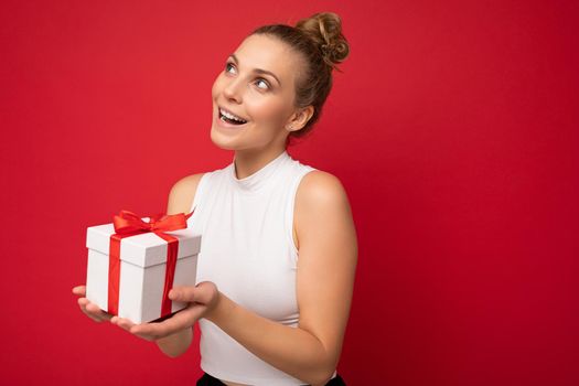 Photo shot of beautiful positive thoughtful young blonde woman isolated over red background wall wearing white top holding gift box and looking to the side and thinking. Empty space