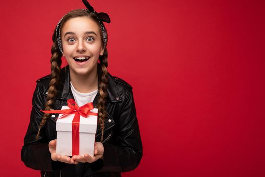 Photo of beautiful happy brunette girl isolated over red background wall wearing white casual t-shirt and stylish black leather jacket holding white gift box with red ribbon and looking at camera. Free space
