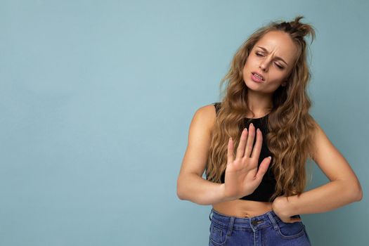 Young dissatisfied sad attractive winsome blonde curly woman with sincere emotions wearing trendy black top isolated over blue background with free space and showing palm. Negative concept.