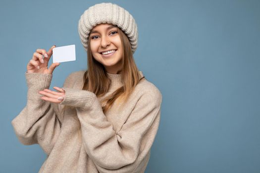 Photo shot of attractive positive smiling young dark blonde woman wearing beige sweater and knitted beige hat isolated over blue background holding and showing credit card looking at camera. copy space