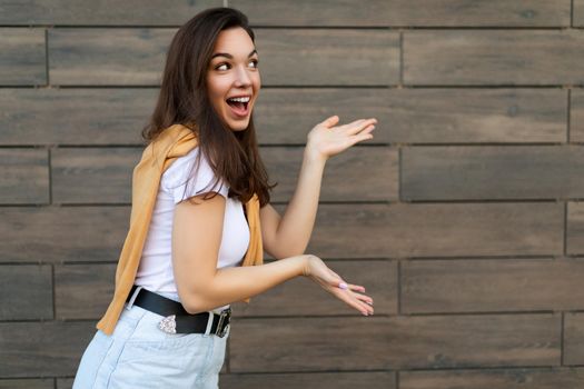 Photo of young emotional happy positive beautiful brunette woman wearing casual clothes standing in the street near the wall and pointing at copy space. Lifestyle concept.