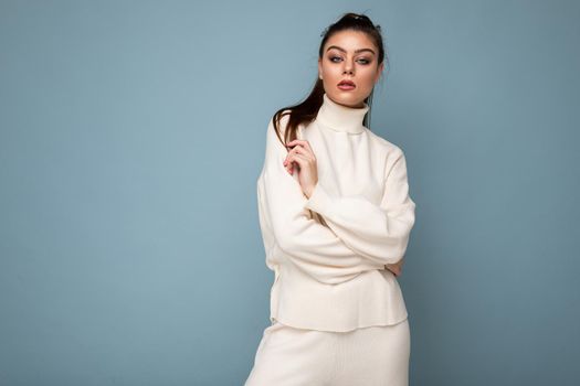 Portrait of a beautiful young brunette woman in white stylish sweater, folded hands in front of her, near the chest, isolated on blue background with copy space.