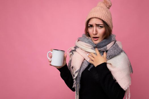 Attractive sick young brunet female person wearing black sweater hat and warm scarf isolated over pink background holding paper mug for mockup drinking hot tea and looking at camera having cold and caughing.