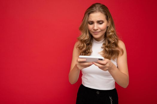 Photo of concentrated beautiful young blonde woman wearing white t-shirt isolated on red background using smartphone and playing phone games looking at gadjet screen.