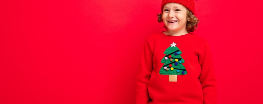 cute blond boy in warm hat and christmas sweater on red background with smile on his face.