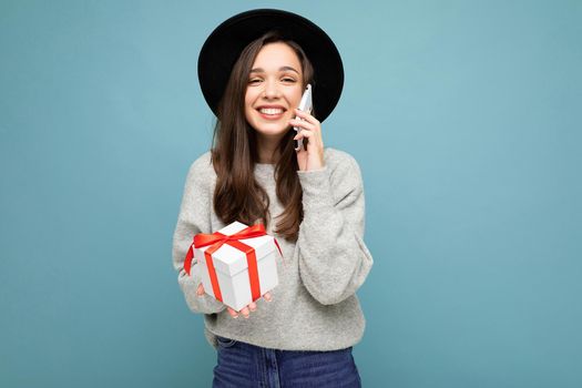 Shot of pretty smiling positive young brunette woman isolated over blue background wall wearing stylish black hat and grey sweater holding gift box talking on smartphone and looking at camera.