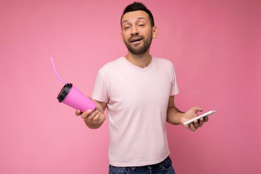 Photo of self-confident handsome young unshaven brunette man with beard wearing everyday light pink t-shirt isolated over pink background holding and using mobile phone surfing on the internet online and drinking beverage looking at camera.