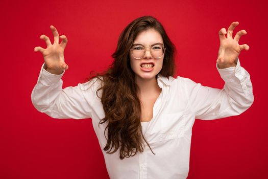 Photo of young emotional attractive brunet lady with sincere emotions wearing casual white shirt and optical glasses isolated on red background with free space and doing claw gesture as cat with agressive and sexy expression.