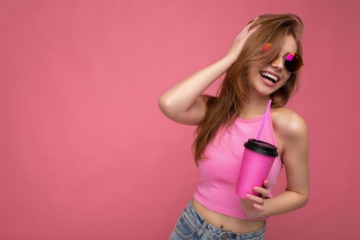 Charming young happy smiling blonde woman wearing stylish clothes isolated over colourful background wall holding paper cup for mockup drinking milkshake looking to the side. empty space
