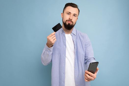 Photo of thoughtful good looking attractive brunet unshaven young man wearing casual blue shirt and white t-shirt isolated over blue background wall holding credit card and using mobile phone making payment looking at camera and thinking.