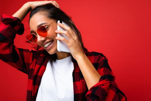 Closeup photo of attractive positive smiling young brunet woman wearing stylish red shirt white t-shirt and red sunglasses isolated over red background communicating on mobile phone looking to the side.