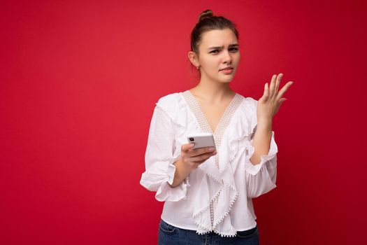 Attractive young upset brunet woman wearing white blouse standing isolated over red background reading news on the internet via phone looking at camera and having questions.