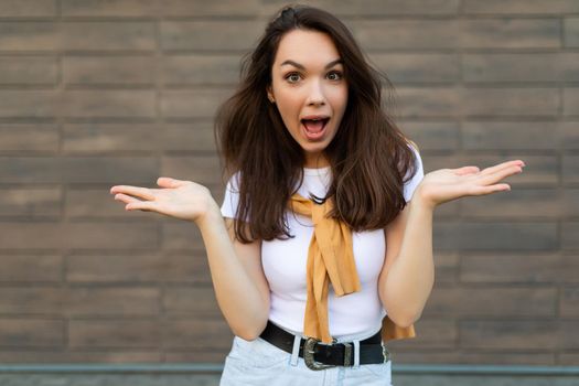Photo of young emotional amazed surprised beautiful brunette woman wearing casual clothes standing in the street near the wall. Lifestyle concept.