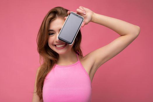 Photo of beautiful smiling young woman good looking wearing casual stylish outfit standing isolated on background with copy space holding smartphone showing phone in hand with empty screen display for mockup looking at camera.