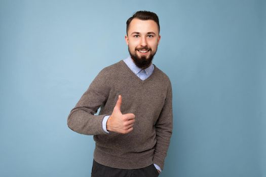 handsome good looking brunet bearded young man wearing grey sweater and blue shirt isolated on pink background with empty space looking at camera and showing thumbs up gesture.